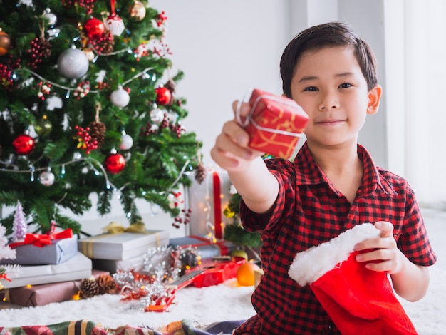 Il ragazzo in una camicia rossa sta tenendo il calzino rosso e felice di festeggiare il Natale con l'albero di Natale