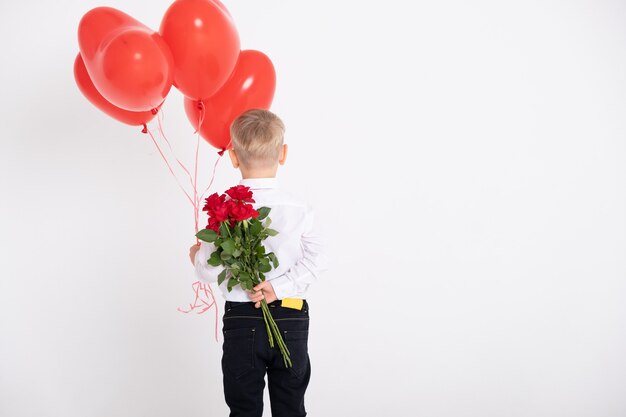 Il ragazzo in vestito tiene il mazzo delle rose e dei palloncini del cuore, sta con le spalle su una priorità bassa bianca.