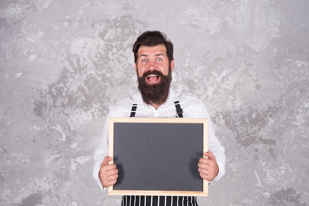 Il ragazzo maturo con barba e baffi è uno chef professionista che dimostra il menu di cucina nella caffetteria sulla lavagna vuota che cucina il cibo