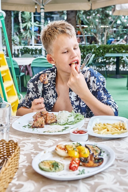 Il ragazzo morde e mastica un pezzo di carne sulla forcella che mangia al tavolo all'aperto nel ristorante