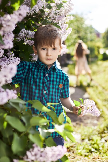 Il ragazzo posa in un cespuglio lilla in primavera. Ritratto romantico di un bambino in fiori alla luce del sole