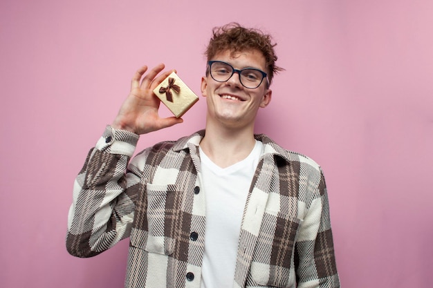 Il ragazzo riccio con gli occhiali mostra una piccola confezione regalo e sorride su uno sfondo rosa un mini regalo per una vacanza e un compleanno