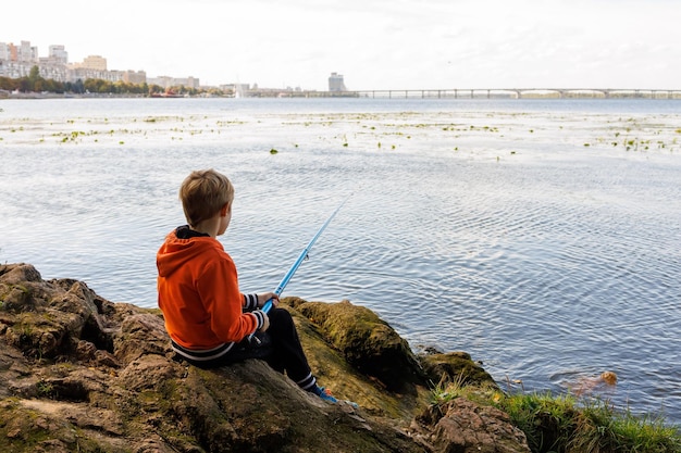 Il ragazzo seduto sui sassi lancia una canna da pesca nel fiume e va a pescare