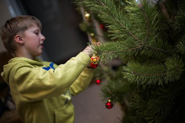 Il ragazzo si aggrappa a una palla rossa su un ramo di un albero di Natale a casa Preparandosi per il Natale a casa