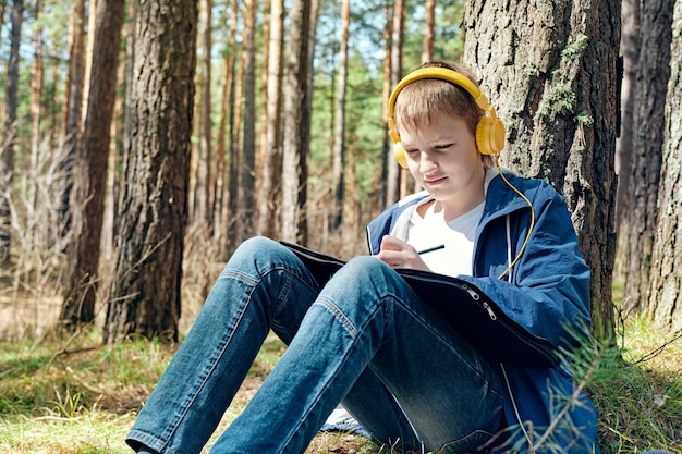 Il ragazzo si siede nella foresta di pini e nella pittura a cielo aperto e ascolta la musica Tempo libero creativo di una giornata di sole