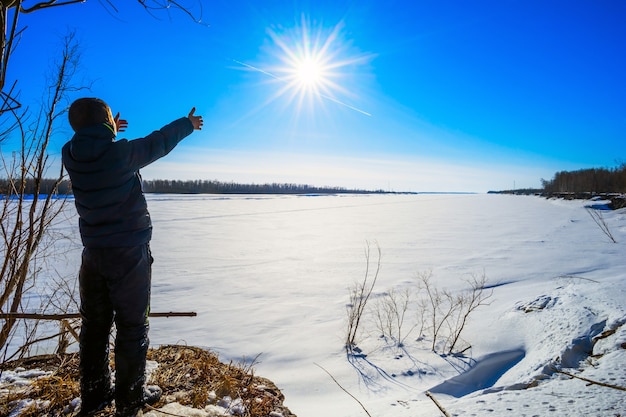 Il ragazzo si trova sulla riva del fiume ghiacciato e tirato a mano al sole.