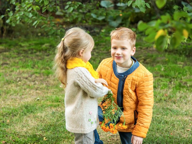 Il ragazzo sorridente allegro in giacca gialla presenta fiori alla bambina in sciarpa gialla in natura