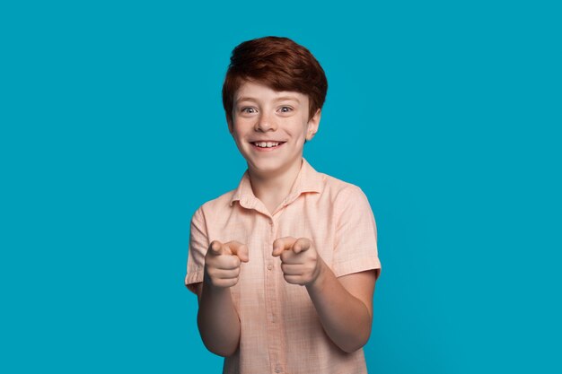Il ragazzo sorridente caucasico sta gesticolando alla macchina fotografica con il dito puntato su una parete blu dello studio con spazio libero