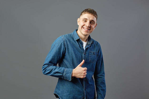 Il ragazzo sorridente vestito con una camicia di jeans tiene il pollice alzato in studio sullo sfondo grigio.