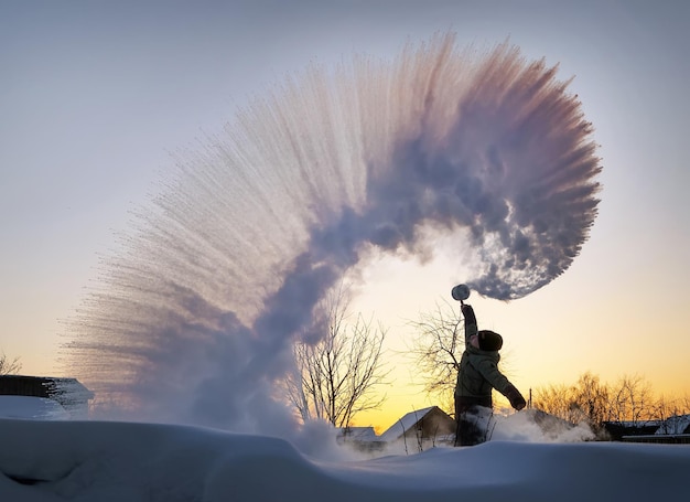 Il ragazzo spruzza acqua bollita da un thermos sopra la sua testa sullo sfondo di un tramonto con tempo gelido. L'effetto del congelamento istantaneo dell'acqua calda in caso di forte gelo. Esperimento di fuochi d'artificio siberiani