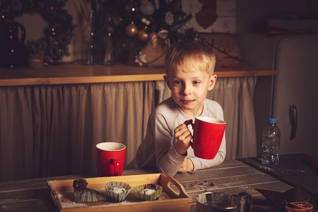 Il ragazzo sta bevendo il tè con i muffin in cucina. Decorazioni natalizie, tradizioni familiari, cibo natalizio, vigilia delle vacanze.