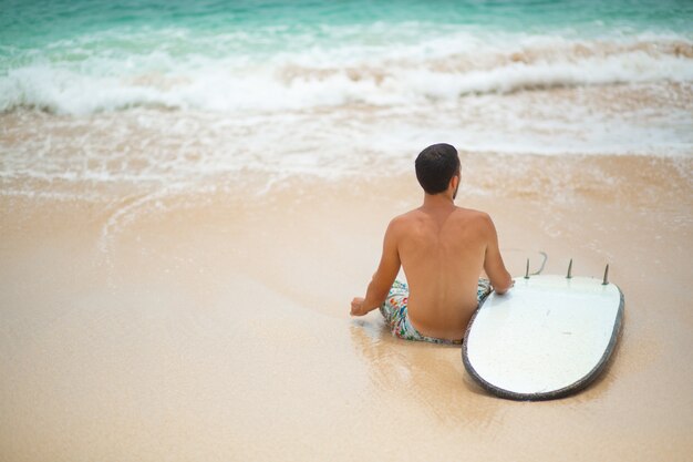 Il ragazzo sta riposando su una spiaggia tropicale di sabbia, dopo aver cavalcato un surf. Stile di vita sano e attivo nella vocazione estiva