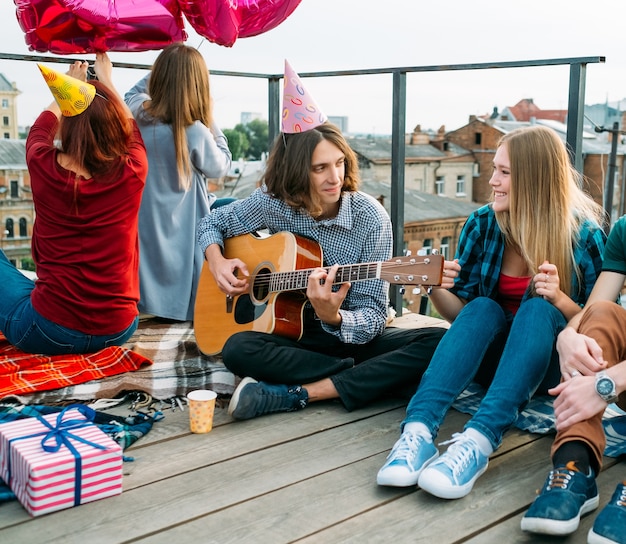 Il ragazzo suona la chitarra e canta una canzone di buon compleanno indossando un cappello da festa. Congratulazioni musicali. Divertimento celebrativo