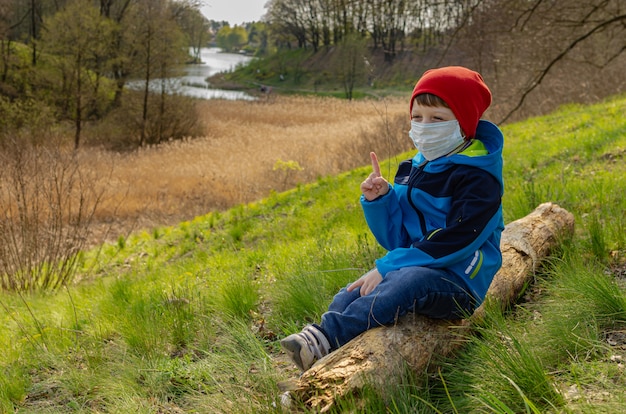 Il ragazzo sveglio in una mascherina medica si siede su una collina su un ceppo e guarda il lago. La famiglia cammina all'aperto con bambini all'inizio della primavera durante la quarantena