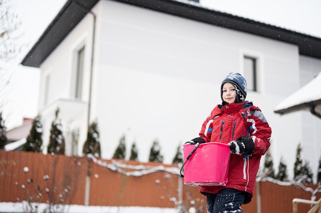 Il ragazzo tiene il secchio con la neve contro la casa in inverno Tempo libero per bambini all'aperto attivo