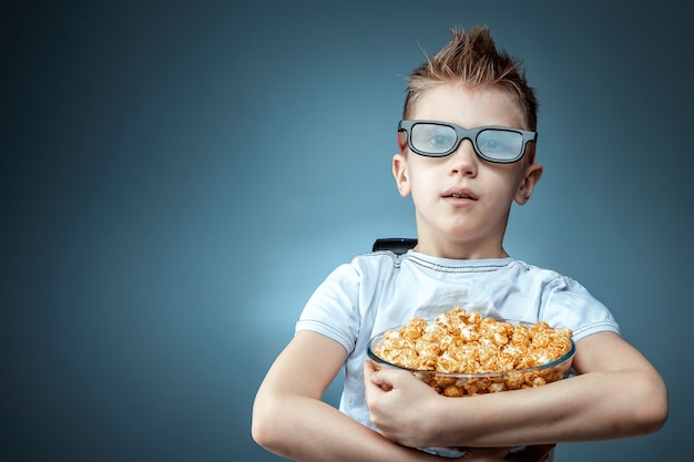 Il ragazzo tiene popcorn nelle sue mani guardando un film con gli occhiali 3D su una parete blu. Il concetto di cinema, film, emozioni, sorpresa, tempo libero.