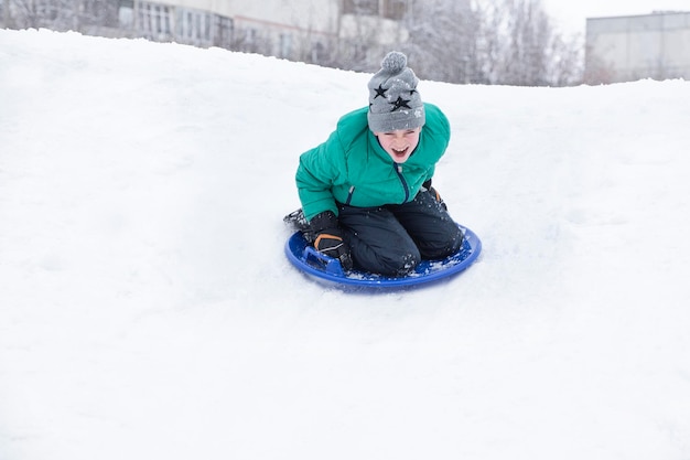 Il ragazzo urlante scivola giù per la collina sul disco da neve Concetto stagionale Giornata invernale