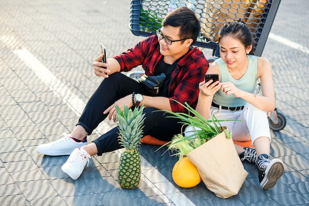 Il ragazzo vietnamita attraente e la ragazza beauiful che si siedono sul parcheggio vicino al centro commerciale e usano i loro smartphone.