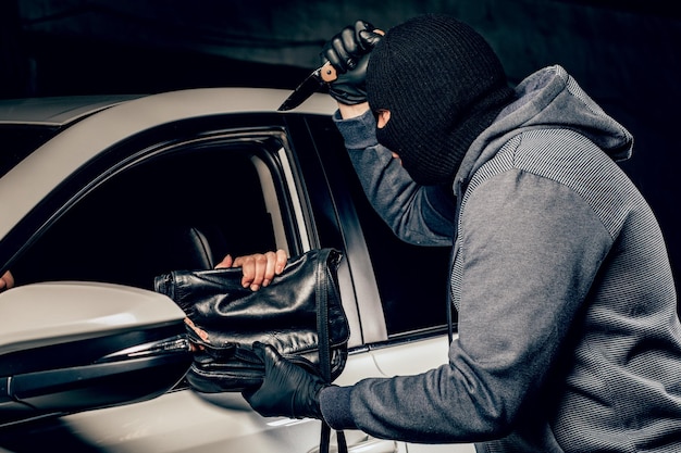Il rapinatore a Balaclava minacciando con un coltello porta via una borsa all'autista di un'auto. Il concetto di criminalità.
