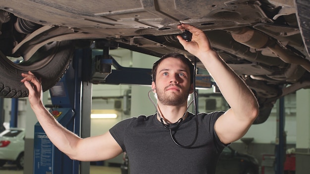 Il riparatore dell'officina dell'automobile è in piedi sotto l'automobile sollevata e i dettagli d'esame.