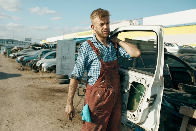 Il riparatore maschio tiene la porta sulla discarica di auto. Rottami di automobili, cianfrusaglie di veicoli, rifiuti di automobili. Trasporti abbandonati, danneggiati e frantumati, cantiere di demolizione