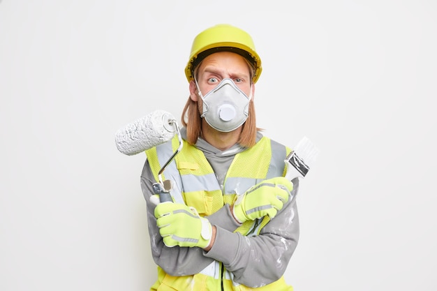 Il riparatore serio indossa la maschera protettiva, il casco e i guanti incrociano le braccia, tiene la spazzola a rullo di vernice stanca delle routine quotidiane durante i lavori di ristrutturazione della casa Uomo costruttore con attrezzatura