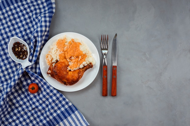 Il riso con salsa al curry con coscia di pollo fritto è servito sul piatto bianco. Asciugamano blu, pepe nella ciotola a forma di pesce, coltello e forchetta decorano sfondo grigio.