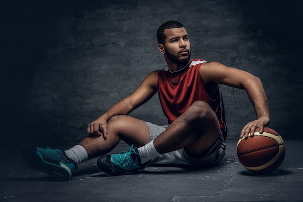 Il ritratto completo del corpo di un giocatore di basket nero si siede su un pavimento e tiene la palla da basket.