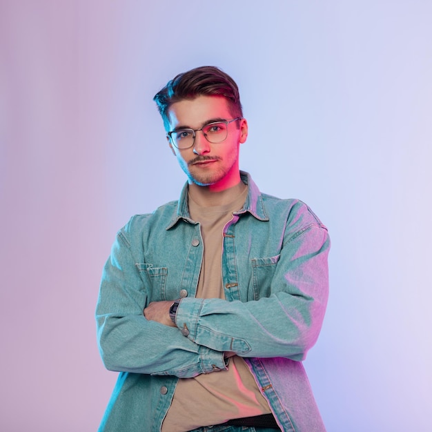 Il ritratto creativo dell'uomo giovane artista bello alla moda con occhiali vintage in abito denim con camicia di jeans sta in studio con luci rosa e blu