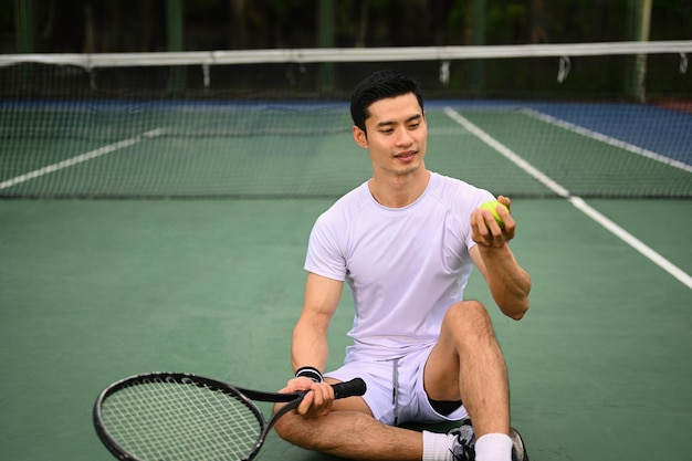 Il ritratto del giocatore di tennis maschio asiatico bello nello sport bianco copre la tenuta della racchetta della palla che si siede sulla corte