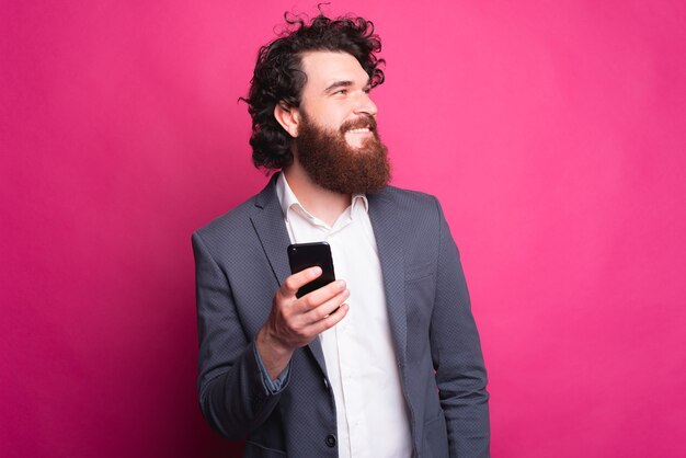 Il ritratto del giovane bello con la barba è vestito che tiene smart phone e distoglie lo sguardo a copyspace