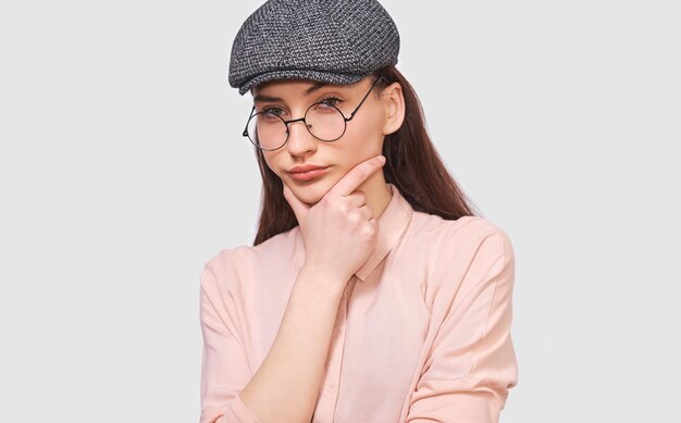 Il ritratto del primo piano della giovane donna pensierosa indossa una camicia casual con occhiali rotondi trasparenti e un berretto grigio guarda seriamente alle pose della fotocamera su sfondo bianco per studio Concetto di persone ed emozioni