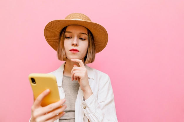 Il ritratto del primo piano della ragazza pensierosa con il cappello su sfondo rosa utilizza lo smartphone con la faccia pensierosa indossa abiti estivi leggeri Signora elegante con la faccia pensierosa isolata Spazio di copia