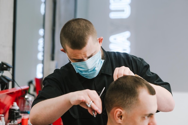 Il ritratto del primo piano di un barbiere professionista con una maschera medica taglia un cliente con le forbici in un coronavirus di quarantena Il parrucchiere utilizza dispositivi di protezione individuale al lavoro Coronavirus
