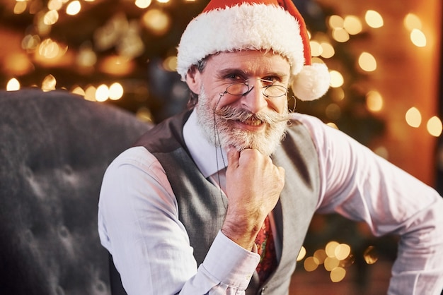 Il ritratto dell'anziano alla moda con capelli grigi e barba in giacca e camicia bianca è nella stanza di natale decorata.