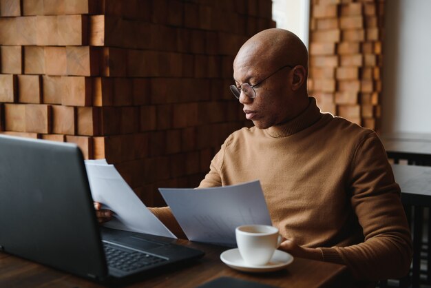 Il ritratto dell'uomo afroamericano sorridente in vetri si siede allo scrittorio in ufficio che lavora al computer portatile