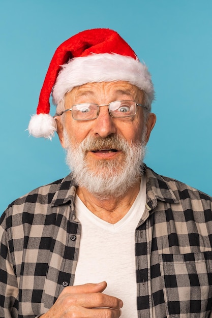 Il ritratto dell'uomo anziano scioccato indossa il cappello di Babbo Natale a bocca aperta isolato su sfondo di colore blu persone anziane e concetto di emozione