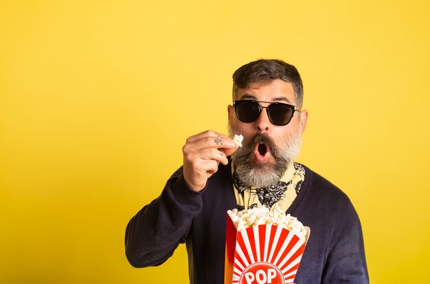 Il ritratto dell'uomo con la barba bianca e gli occhiali da sole che mangiano il popcorn hanno stupito la televisione di sorveglianza su fondo giallo.