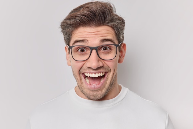 Il ritratto dell'uomo dai capelli scuri felice sorpreso fissa con gli occhi pieni di gioia ride positivamente indossa occhiali trasparenti e maglione casual isolato su sfondo bianco Concetto di reazioni umane