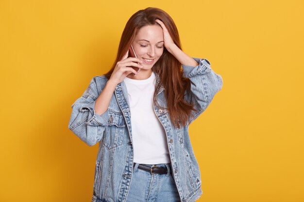 Il ritratto della bella donna piacevole veste la giacca alla moda del denim e la camicia casuale bianca, tenendo lo smartphone