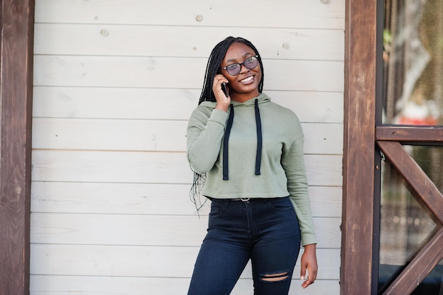 Il ritratto della città di una giovane donna dalla pelle scura positiva che indossa una felpa con cappuccio verde e occhiali da vista parla sul telefono cellulare