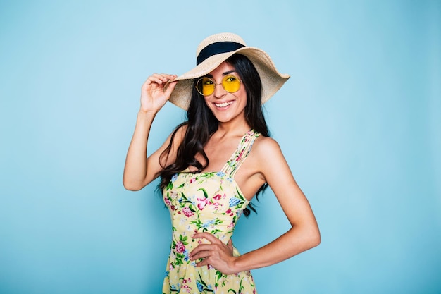 Il ritratto della donna castana sveglia dell'estate in occhiali da sole del cappello e la ragazza alla moda del vestito variopinto si divertono e posano su priorità bassa blu