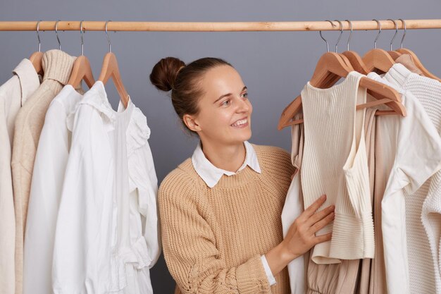 Il ritratto della donna caucasica positiva sorridente attraente che sta vicino ai vestiti appende sullo scaffale che sceglie la camicia da una nuova raccolta che sorride godendo lo shopping