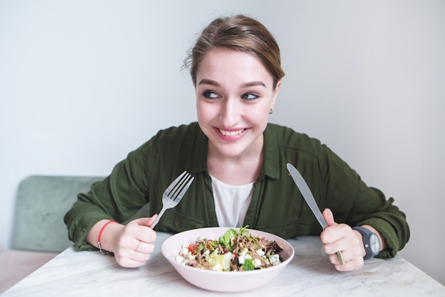 Il ritratto della donna divertente con la coltelleria in sue mani si avvicina al piatto di insalata.