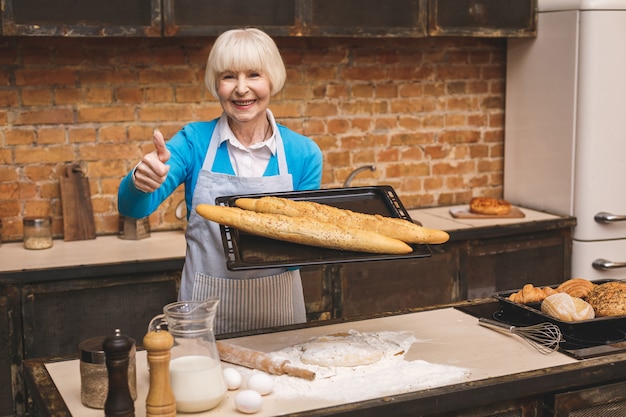 Il ritratto della donna invecchiata senior felice sorridente attraente sta cucinando sulla cucina. Nonna che produce una cottura saporita. Pollice su.