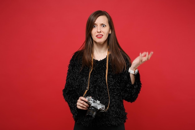 Il ritratto della giovane donna perplessa in maglione di pelliccia nera tiene la retro macchina fotografica della foto dell'annata isolata sul fondo rosso luminoso della parete in studio. Persone sincere emozioni, concetto di stile di vita. Mock up copia spazio.