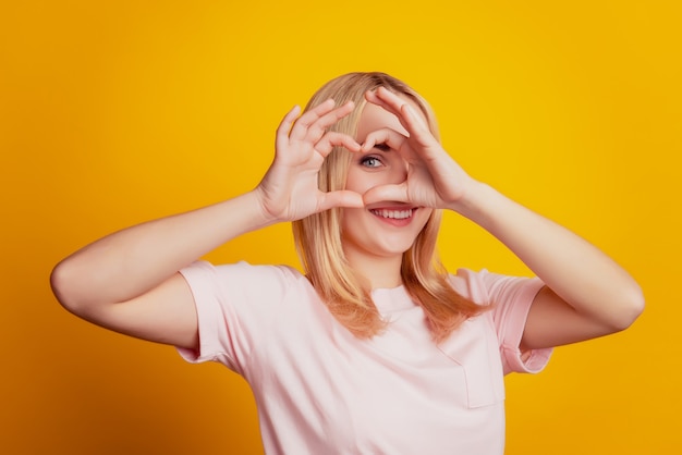 Il ritratto della ragazza affettuosa adorabile divertente mostra l'occhio della copertura di gesto del cuore su fondo giallo