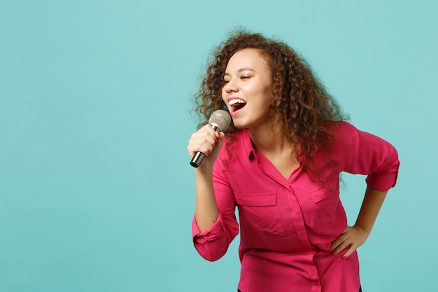 Il ritratto della ragazza africana divertente in vestiti casuali che balla, canta la canzone in microfono isolato sul fondo blu della parete del turchese in studio. Persone sincere emozioni, concetto di stile di vita. Mock up copia spazio.