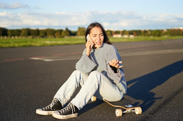 Il ritratto della ragazza asiatica si siede sul suo longboard pattinando sullo skateboard e parlando al telefono cellulare havin