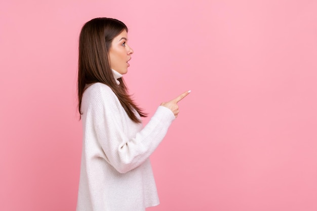 Il ritratto della vista laterale della ragazza stupita punta da parte con il dito, mostra lo spazio vuoto per l'idea commerciale, indossando un maglione bianco in stile casual. Studio indoor girato isolato su sfondo rosa.